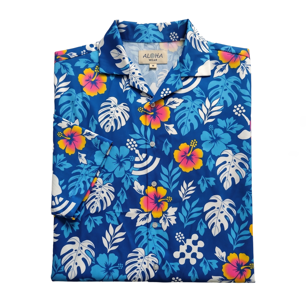 A folded short-sleeve button-up shirt with a vibrant tropical floral print on a royal blue background. The pattern features white monstera leaves, blue palm fronds, and hibiscus flowers in pink, yellow, and orange. custom sublimated with company logo